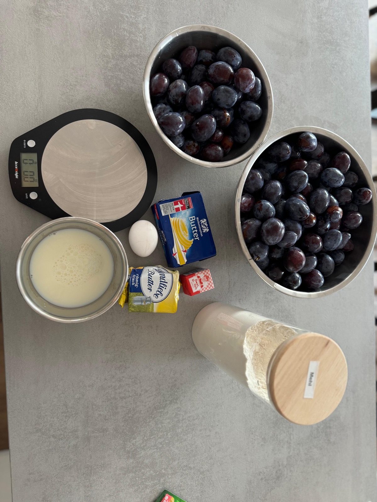 Zutaten für den Hefeteig: Mehl, Butter, Milch, Ei und Hefe neben Schüsseln voller Zwetschgen